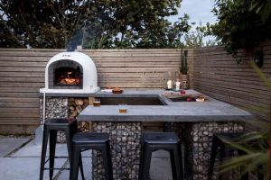 Outdoor kitchen with benchtop, sink, seating and oven in a fenced backyard.
