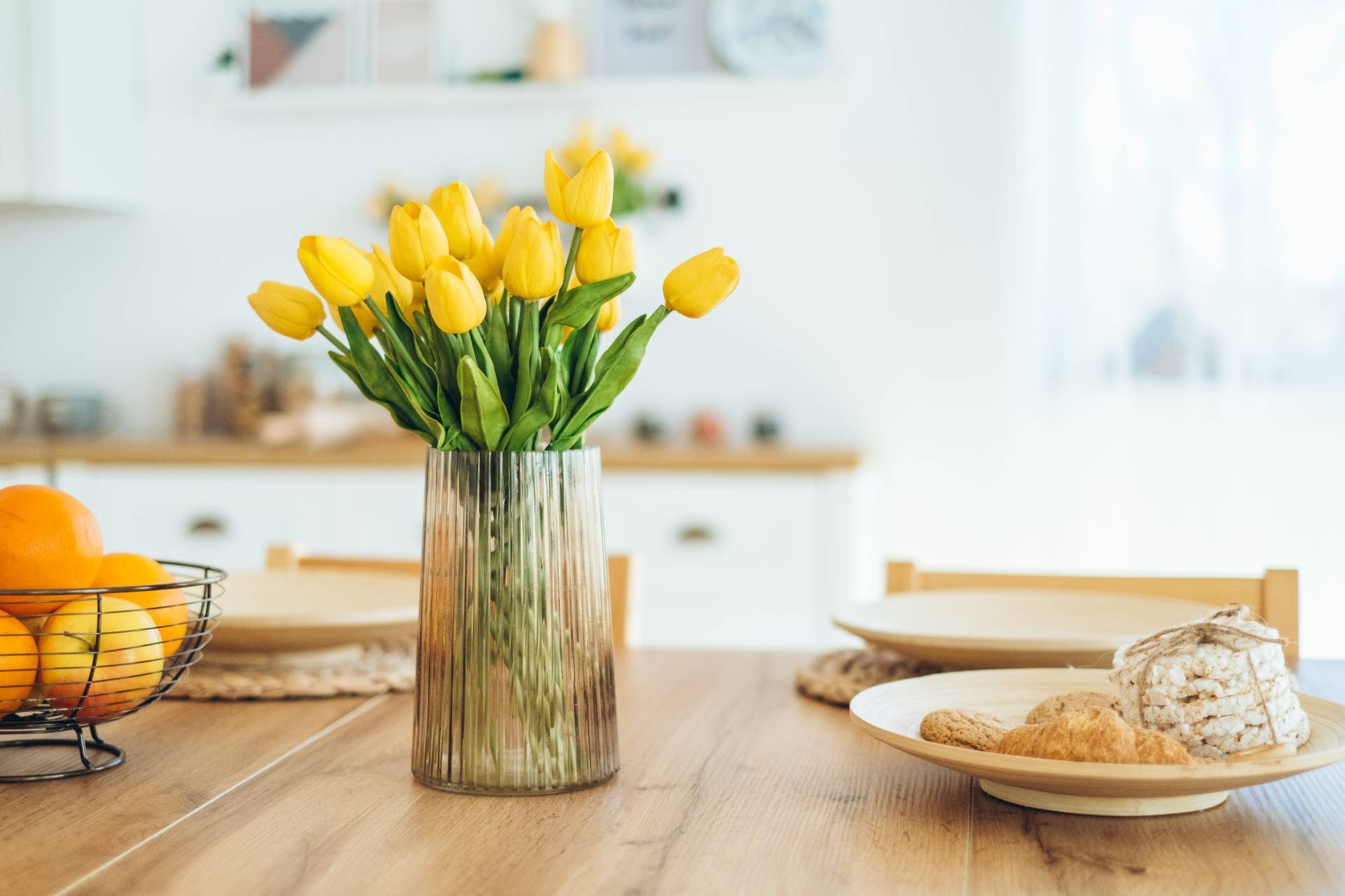 a vase of yellow tulips on a wooden dining table with plates of snacks and a bowl of oranges