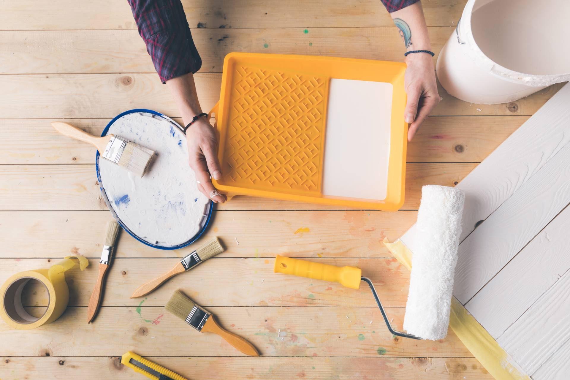DIY house painting tools on wooden surface with brushes, roller, tape, tray and white paint bucket