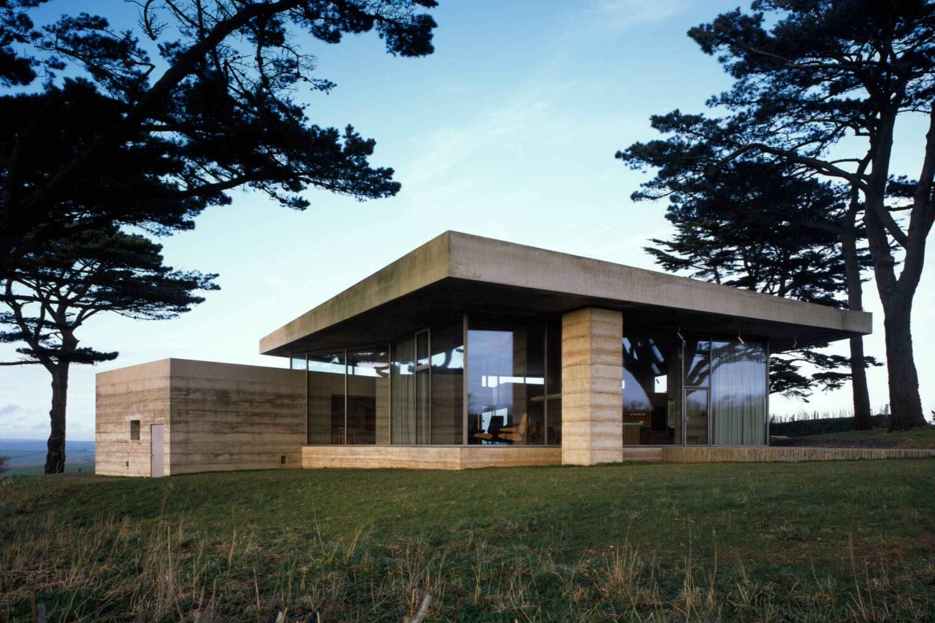 Modern concrete house with large glass walls set among tall trees on a grassy hill